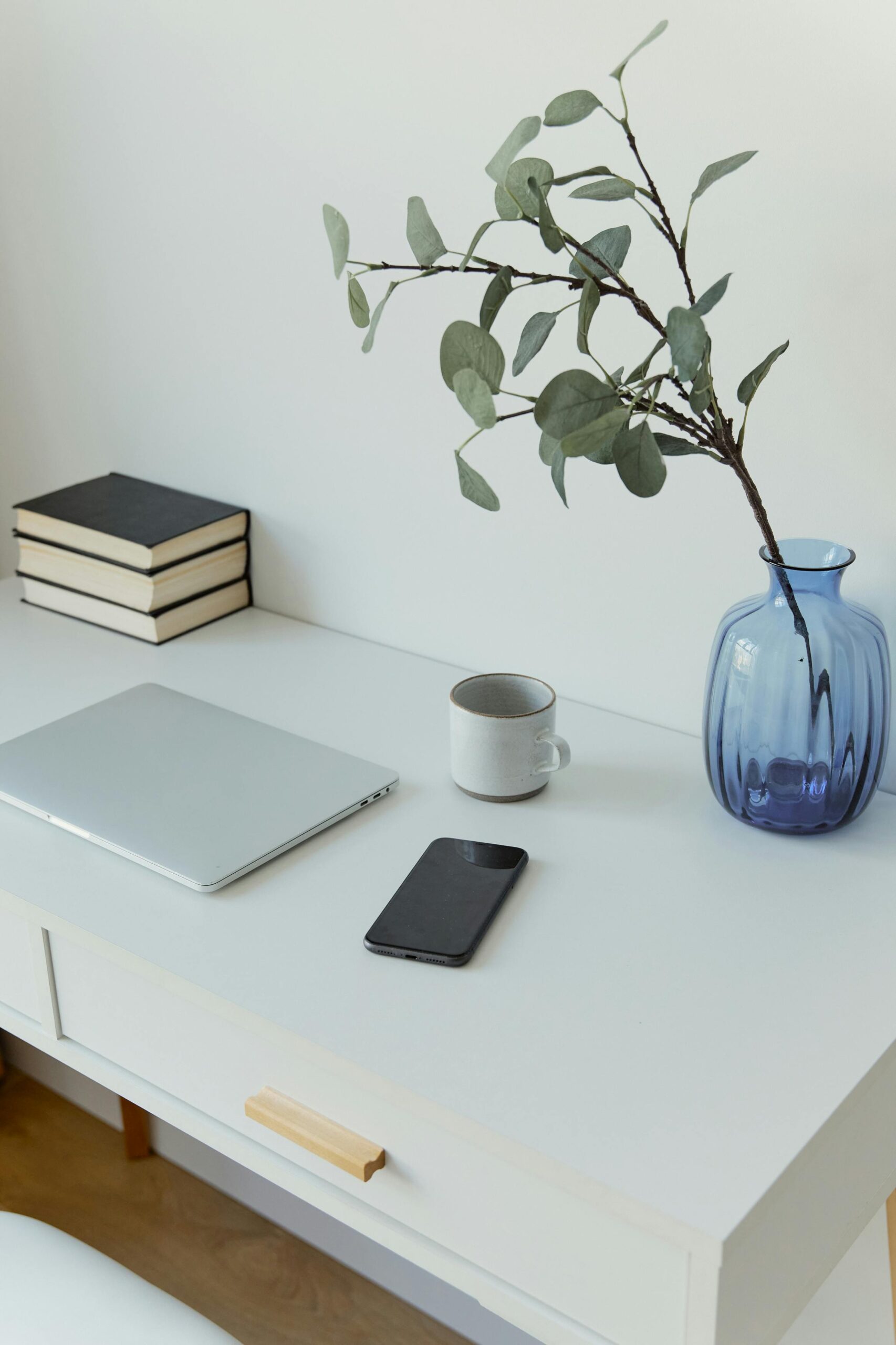 A clean and minimalist workspace featuring a laptop, smartphone, and decorative vase on a white desk.
